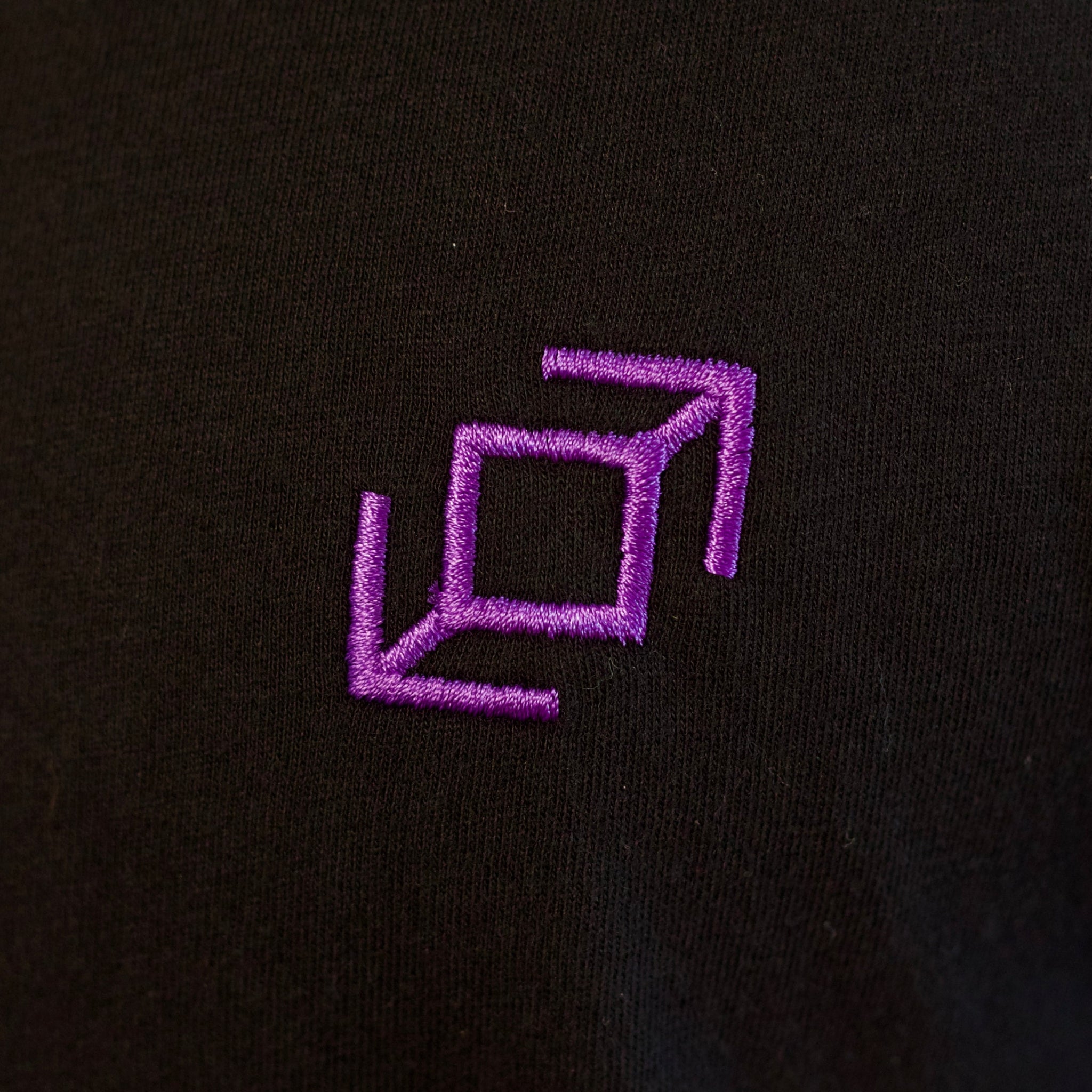 A close up of the purple embroidery of a whip symbol on a black cotton fabric